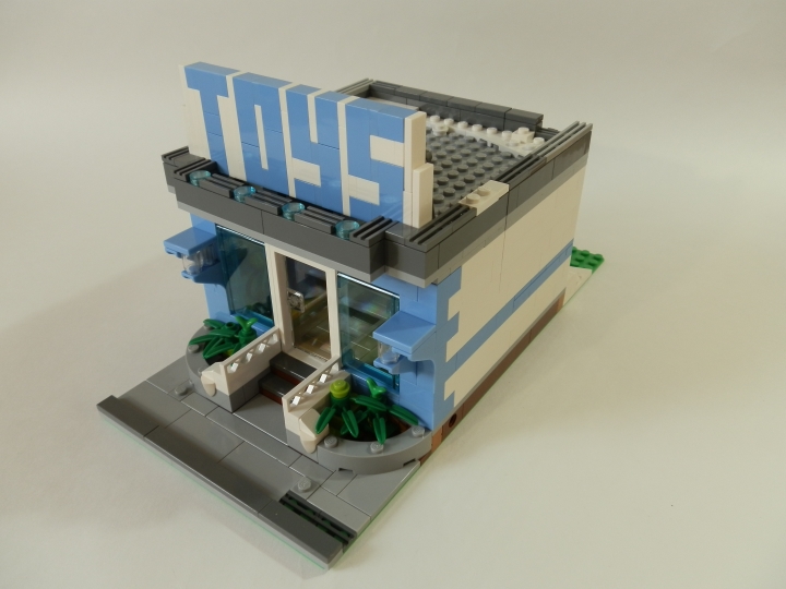 LEGO MOC - New Year's Brick 2014 - Магазин игрушек.: Теперь осмотрим само строение, не обращая внимание на народ.<br />
Клумбы со стороны фасада сильно скрадывают часть тротуара, но почему бы и нет? Сам фасад состоит в основном из стекла, что позволяет осмотреть все внутренности с улицы. Выше - крыша и вывеска на ней. Огромные буквы не оставят сомнений о товарах в этом магазине.